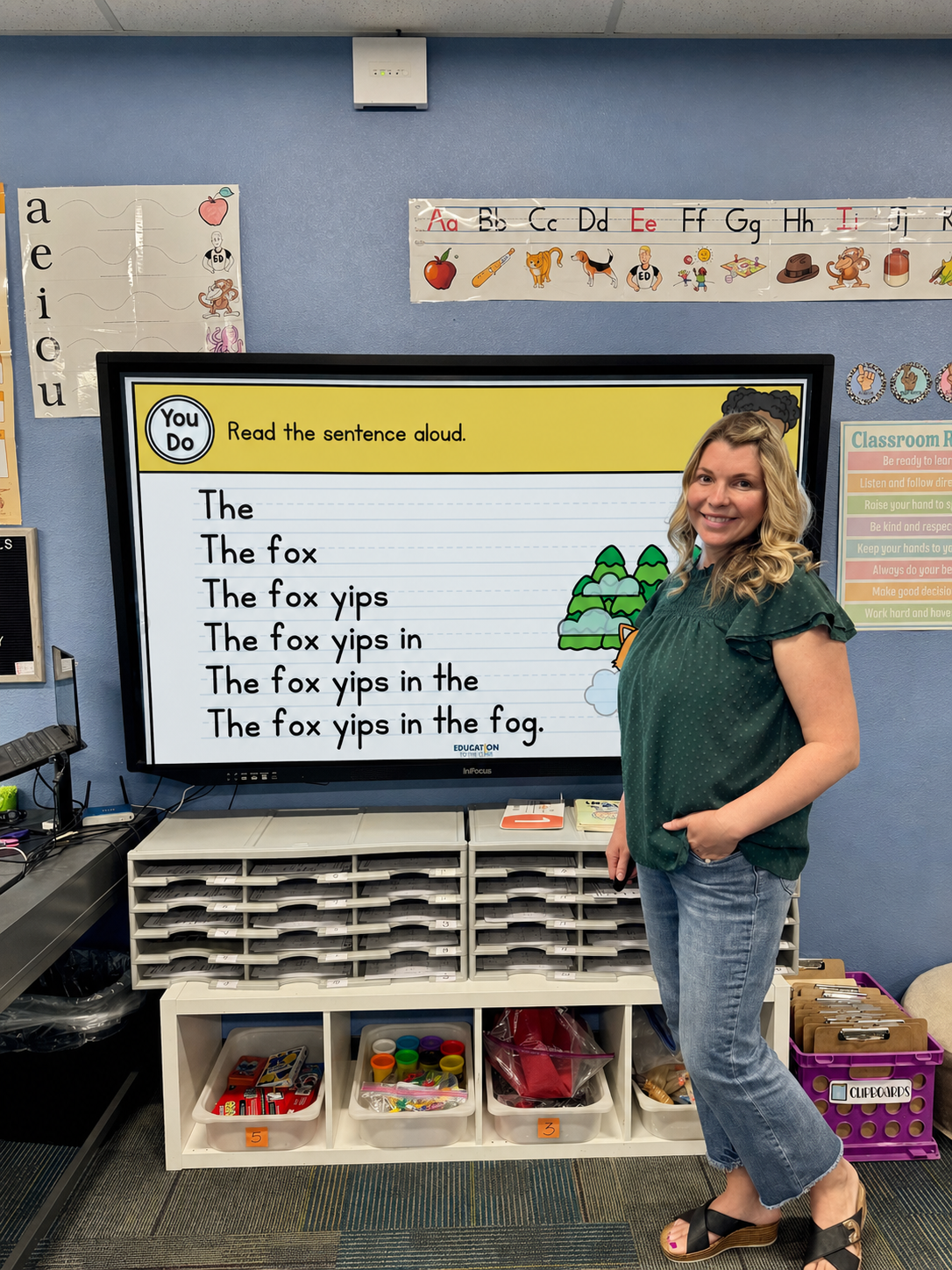 Teacher stands beside a large interactive screen with a read-aloud sentence about 'The fox' for a phonics lesson.